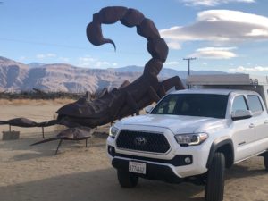 A large steel sculpture of a scorpion behind a white pickup truck of the same size.